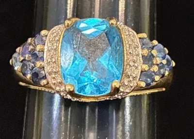 Sale Photo Thumbnail #119: Gold cocktail ring featuring a large oval cut blue topaz center stone flanked by pave diamond and sapphire accents.