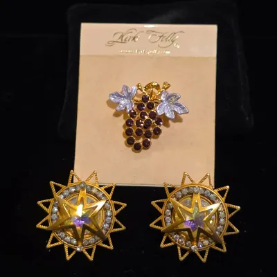 Sale Photo Thumbnail #122: Kirks Folly grape cluster brooch with purple rhinestones on its original card, alongside a pair of gold-tone starburst clip-on earrings with iridescent crystal centers.