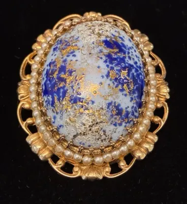 Sale Photo Thumbnail #85: Vintage gold-tone brooch featuring an oval blue and gold splatter art glass cabochon framed by a faux pearl trim and openwork filigree edge.