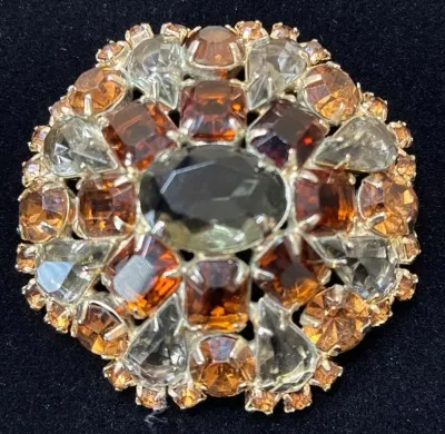 Sale Photo Thumbnail #80: Vintage circular rhinestone brooch featuring amber, smoke grey, and clear prong-set stones in a silvertone setting.