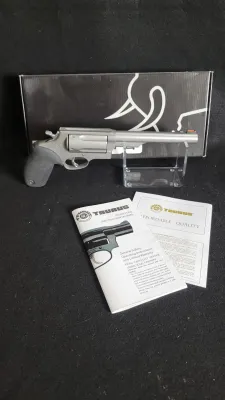 Taurus stainless steel Judge revolver with textured black rubber grips, shown on a display stand with its original box and instruction...