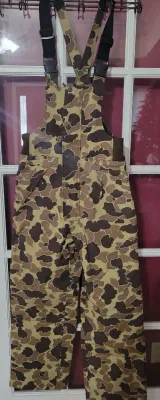 Vintage duck camouflage hunting bib overalls with adjustable shoulder straps and side stretch panels, shown hanging on a door.