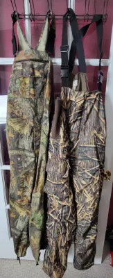 Two pairs of camouflage hunting bib overalls hanging from a rack, featuring a Liberty brand pair on the left and a Mossy Oak Shadow Grass...