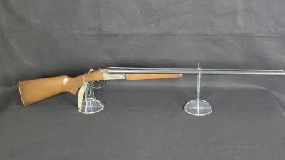 Vintage side-by-side double-barrel shotgun with a wooden stock and forend, displayed on clear acrylic stands against a dark backdrop.