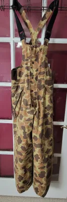 Vintage duck camouflage hunting bib overalls featuring adjustable shoulder straps, button-front closure, and side zip openings.