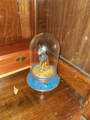 Sale Photo Thumbnail #111: Egyptian-revival Bastet cat figurine enclosed in a glass cloche on a blue hieroglyphic-patterned base.