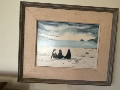 Sale Photo Thumbnail #299: Framed watercolor painting depicting three nuns seated on a sandy beach looking out at the ocean under a cloudy sky.