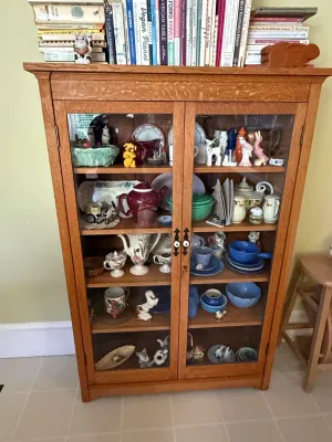 Sale Photo Thumbnail #386: Mission-style quarter-sawn oak display cabinet filled with vintage ceramics, figurines, and glassware, topped with a collection of cookbooks.
