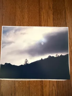 Sale Photo Thumbnail #268: Unframed landscape photograph print depicting a moonlit or sunlit overcast sky over silhouetted hills and trees, resting on a wood floor surface.
