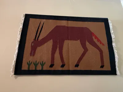 Sale Photo Thumbnail #193: Hand-woven wool tapestry wall hanging depicting a stylized horned antelope or gazelle grazing on a tan background, framed by a dark border with fringe.