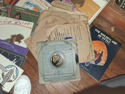 Sale Photo Thumbnail #126: A scattered collection of vintage vinyl records, antique 78 RPM shellac discs in paper sleeves, and art books resting on a wooden floor.