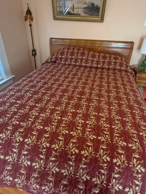 Sale Photo Thumbnail #265: Traditional wooden full or queen bed frame topped with a burgundy and gold vine-patterned bedspread. The bedroom setup includes a framed landscape painting overhead and a vintage floor lamp alongside.