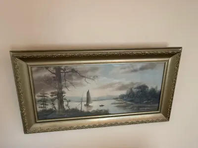 Sale Photo Thumbnail #238: Framed horizontal landscape painting depicting a sailboat on a serene lake surrounded by trees, housed in an ornate molded metallic frame.