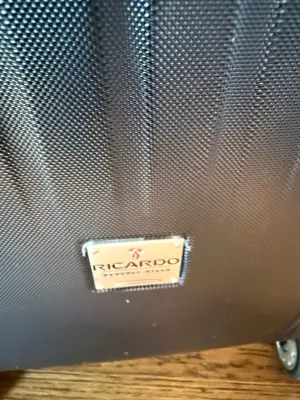 Sale Photo Thumbnail #228: Close-up of a Ricardo Beverly Hills brand metal plaque on a dark gray woven fabric suitcase.