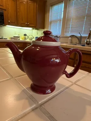 Sale Photo Thumbnail #468: Glossy burgundy ceramic teapot featuring a white inner lid rim and a pediment base, displayed on a tiled kitchen counter.