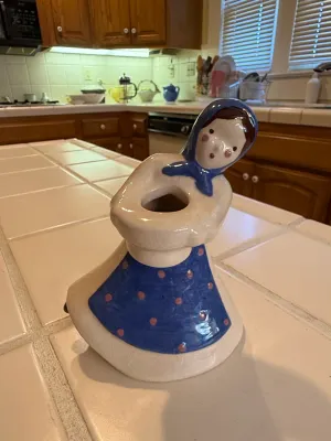 Sale Photo Thumbnail #498: Vintage ceramic figurine planter shaped like a woman wearing a blue headscarf and polka-dot apron, sitting on a tiled countertop.