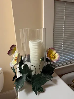 Sale Photo Thumbnail #400: Glass hurricane candle holder featuring a floral base with faux leaves and purple and yellow flowers, holding a textured white pillar candle.