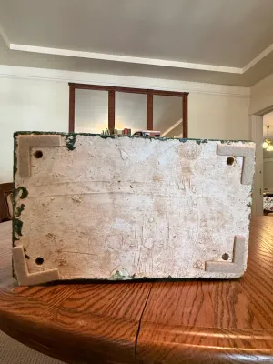 Sale Photo Thumbnail #186: Underside of a heavy composition or concrete decorative object showing corner felt pads and drainage or mounting holes, displayed on a wooden dining table.