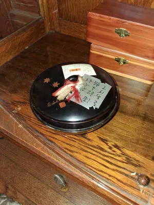Sale Photo Thumbnail #76: Black lacquer round lidded box with Japanese calligraphy paper and a decorative tassel resting on top, displayed alongside small wooden chests.
