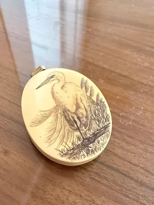 Sale Photo Thumbnail #270: Oval scrimshaw pendant featuring a detailed etch of an egret or heron in a marshland setting, finished with a metal bail.