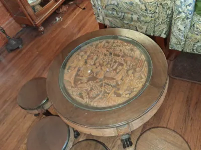 Sale Photo Thumbnail #116: Vintage Asian carved wood round coffee table with a glass insert over a detailed narrative scene, accompanied by matching tuck-away stools.