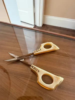 Sale Photo Thumbnail #281: Vintage ornamental scissors featuring amber and mother-of-pearl style acrylic handles with gold-tone decorative filigree accents, displayed on a glass table.