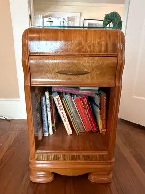 Sale Photo Thumbnail #251: Art Deco waterfall nightstand featuring a top drawer, glass top protector, and an open lower shelf filled with various books.