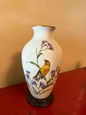 Sale Photo Thumbnail #131: White porcelain vase featuring a hand-painted yellow bird among purple flowers, accented with a gilt rim and a dark wooden base.