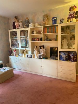 Large white composite wall unit with glass-front cabinets, shelves, and lower drawers, filled with a collection of porcelain dolls,...