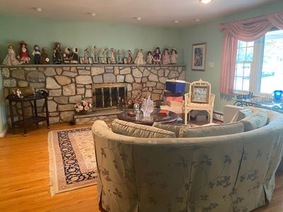 Living room setup featuring a curved floral sofa, a stone fireplace display of vintage porcelain dolls and figurines, an area rug, and...
