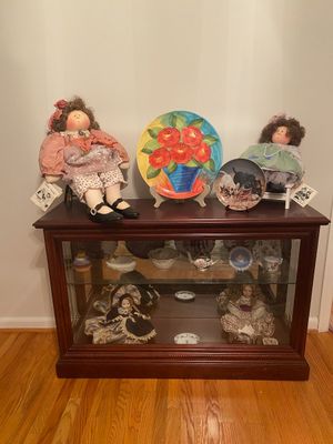 A wooden glass-front display cabinet filled with porcelain dolls and china, topped with soft-sculpture Little Souls dolls and decorative...