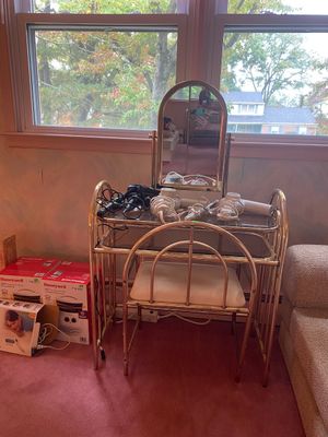 Vintage 1980s brass-finished vanity set with arched mirror, glass top, matching chair, and assorted hair styling tools.