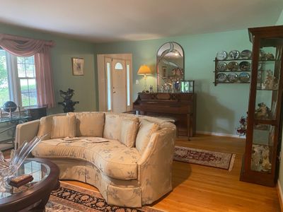 Living room setup featuring a floral curved sectional sofa, a dark wood display cabinet, a piano with bench, and wall-mounted decorative...