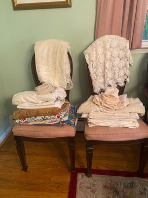 Stacks of assorted vintage linens, including lace tablecloths, napkins, and floral textiles, displayed on a pair of pink upholstered...
