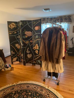 Vintage fur coats hanging on a rolling rack in front of a four-panel decorative folding room divider screen with floral motifs.
