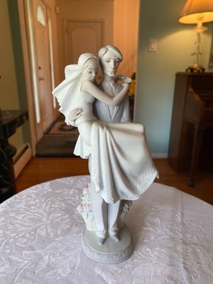 Lladro-style porcelain figurine depicting a groom carrying his bride, featuring a matte finish with delicate lace and floral accents.