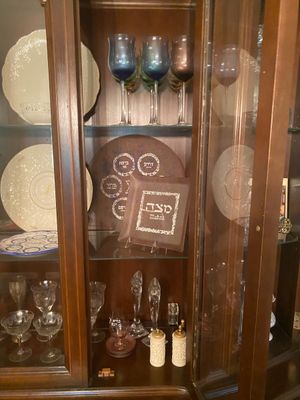Collection of Judaica, glassware, and serving pieces displayed inside a wooden china cabinet, featuring Passover Seder and Matzo plates.