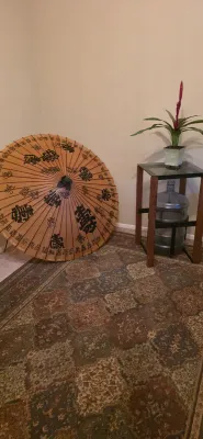 Sale Photo Thumbnail #65: Large Asian oil-paper parasol with calligraphy detailing resting in a room corner, alongside a glass-top accent table housing a potted plant and a decorative glass jug.
