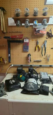 Sale Photo Thumbnail #28: Workshop workbench and pegboard filled with assorted hand tools, power tool battery chargers, knee pads, and a custom wooden jar rack for small hardware.