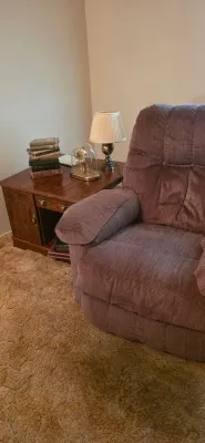 Sale Photo Thumbnail #23: Plush mauve fabric recliner paired with a dark wood side table holding a brass table lamp, glass-dome anniversary clock, and a stack of vintage hardcover books.