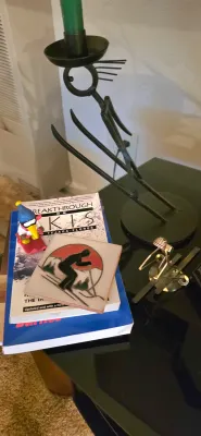 Sale Photo Thumbnail #16: Collection of skiing-themed decor and books, featuring a wrought iron skier candlestick holder, miniature skier figurine, ceramic tile coaster, and 'Breakthrough on Skis' paperback book.