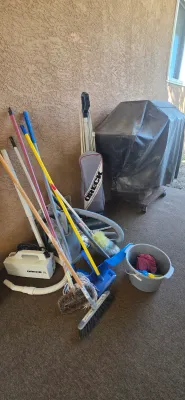 Sale Photo Thumbnail #54: Assorted household cleaning supplies including Oreck XL vacuums, brooms, mops, dusters, and a plastic bucket, displayed outdoors next to a covered outdoor grill.