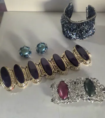 Sale Photo Thumbnail #11: Collection of vintage costume jewelry, including a gold-tone panel bracelet with dark purple stones, a filigree hinged bangle, a beaded wide cuff, and blue clip-on earrings.