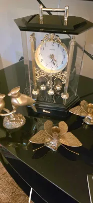 Sale Photo Thumbnail #2: Brass and glass Miyabi quartz anniversary clock with a rotating pendulum, flanked by brass bird figurines and butterfly-shaped candle holders.