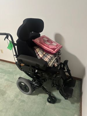 Sale Photo Thumbnail #42: Black motorized power wheelchair with adjustable headrest, leg rests, and padded armrests, shown with blankets and a throw pillow on the seat.