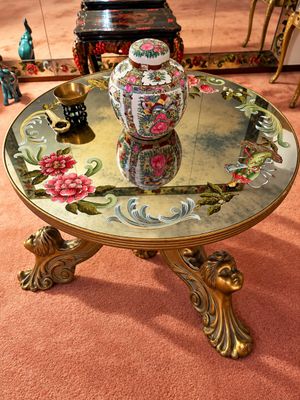 Sale Photo Thumbnail #5: Round coffee table featuring an ornate carved cherub-head base and a painted mirror top decorated with floral designs. Displayed on top are a Rose Medallion porcelain ginger jar and small brass decorative items.