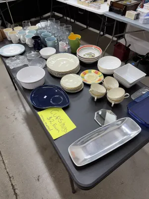 Sale Photo Thumbnail #22: Assorted tableware and kitchenware including ceramic mugs, glassware, serving platters, stacks of dinner plates, mixing bowls, and a metal tray.