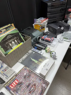 Sale Photo Thumbnail #29: Collection of mixed household electronics and media, including vinyl records, a slide projector, cassette tapes, remote controls, and vintage magazines.