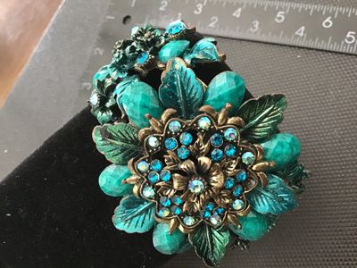 Sale Photo Thumbnail #142: An ornate vintage brass-tone hinged cuff bracelet featuring an elaborate floral design set with sparkling blue rhinestones and green/turquoise-colored cabochons and painted leaf accents. Features a spring hinge closure mechanism. Shows minor finish wear c