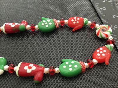 Sale Photo Thumbnail #74: A festive Christmas-themed beaded necklace featuring lampwork glass beads shaped like red and green mittens, holiday boots, and peppermints, interspersed with spacer beads. Measures approximately 28 to 30 inches long with a gold-tone toggle clasp. In good